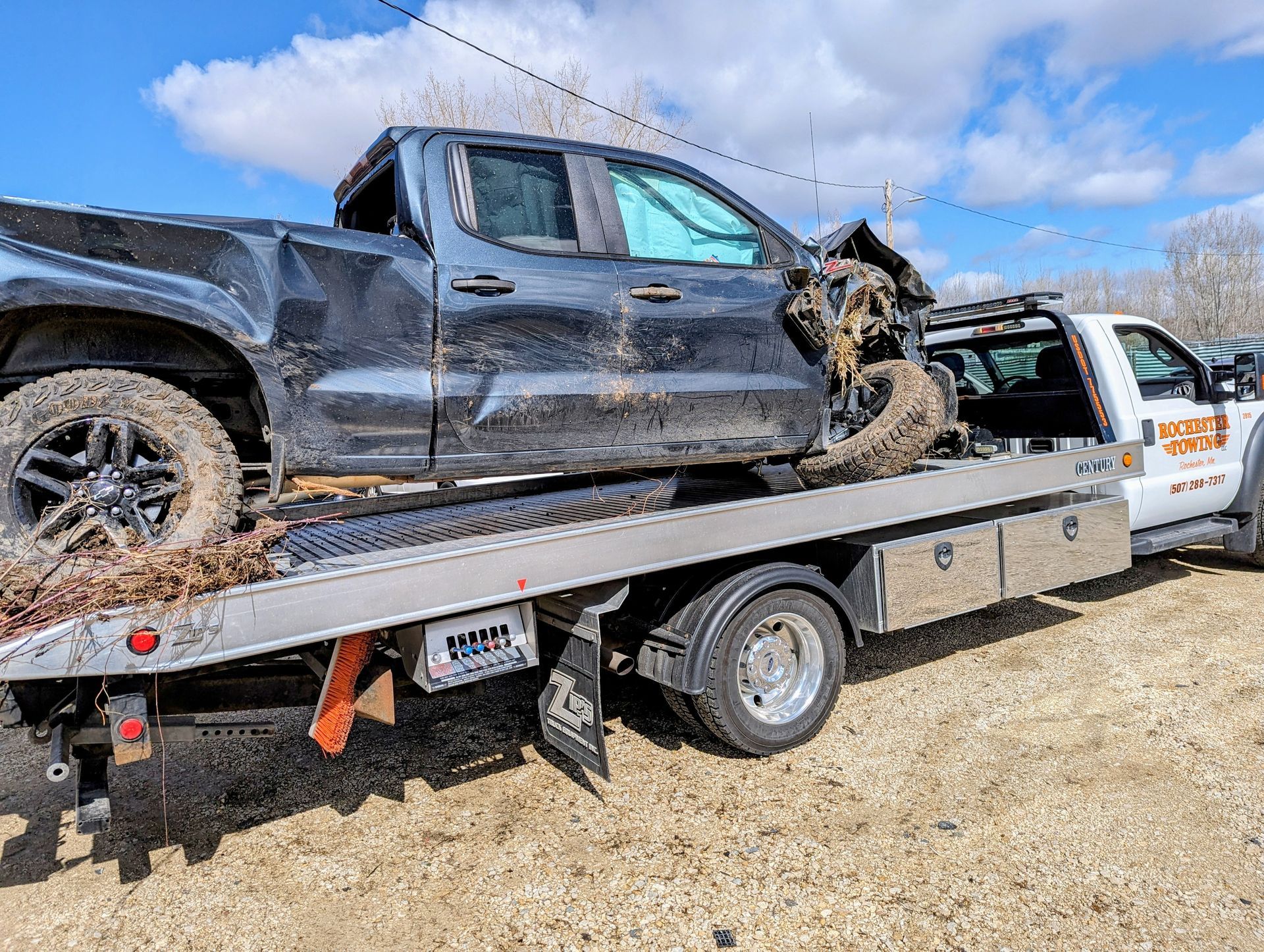 Damaged silver sedan on a tow truck, crumpled rear end, sunny day, next to green grass.