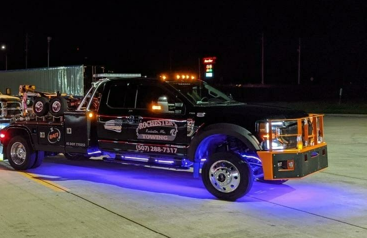 Black tow truck with blue underglow lights parked at night.