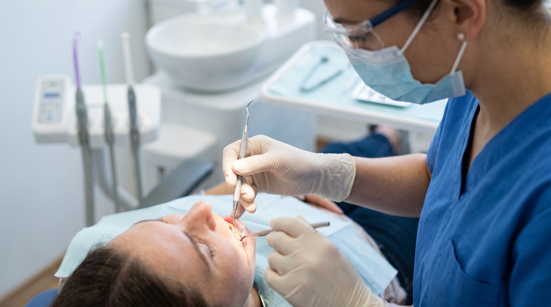 Dentist examining a patient’s mouth with a dental mirror, wearing blue scrubs and a mask in a clinic