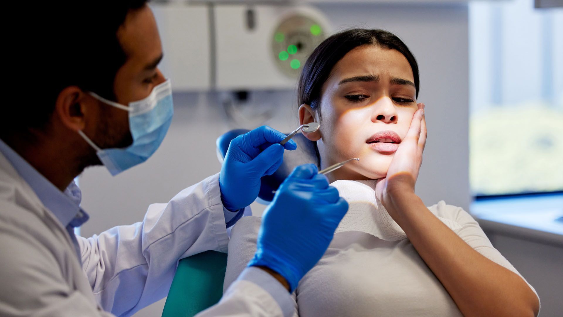 Dentist examining a woman's jaw. The woman has a worried expression, holding her face. Dentist wears a mask and gloves.