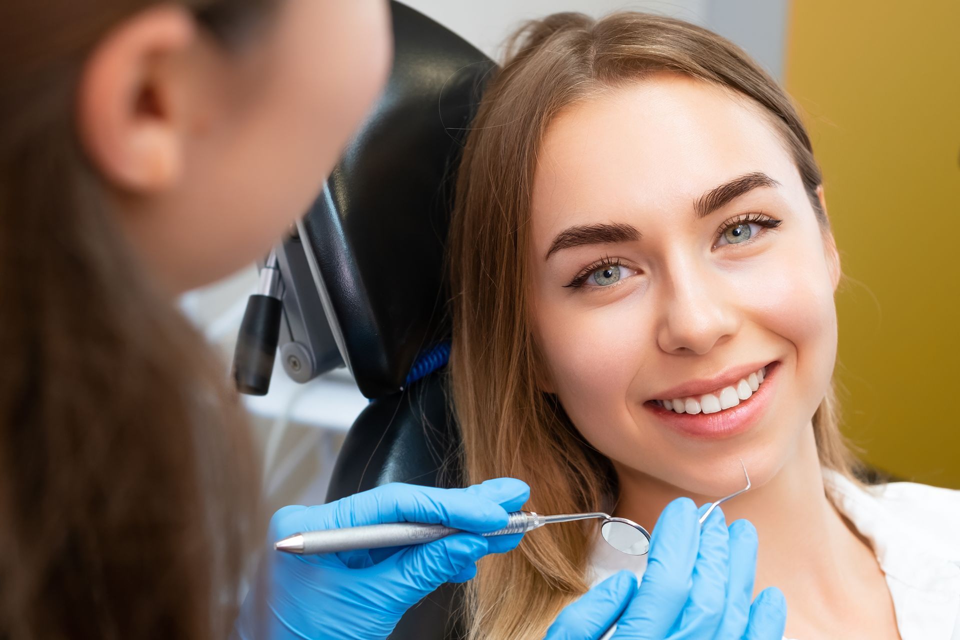 A person in a dental chair smiling while a clinician in blue gloves uses a dental mirror to examine their teeth.