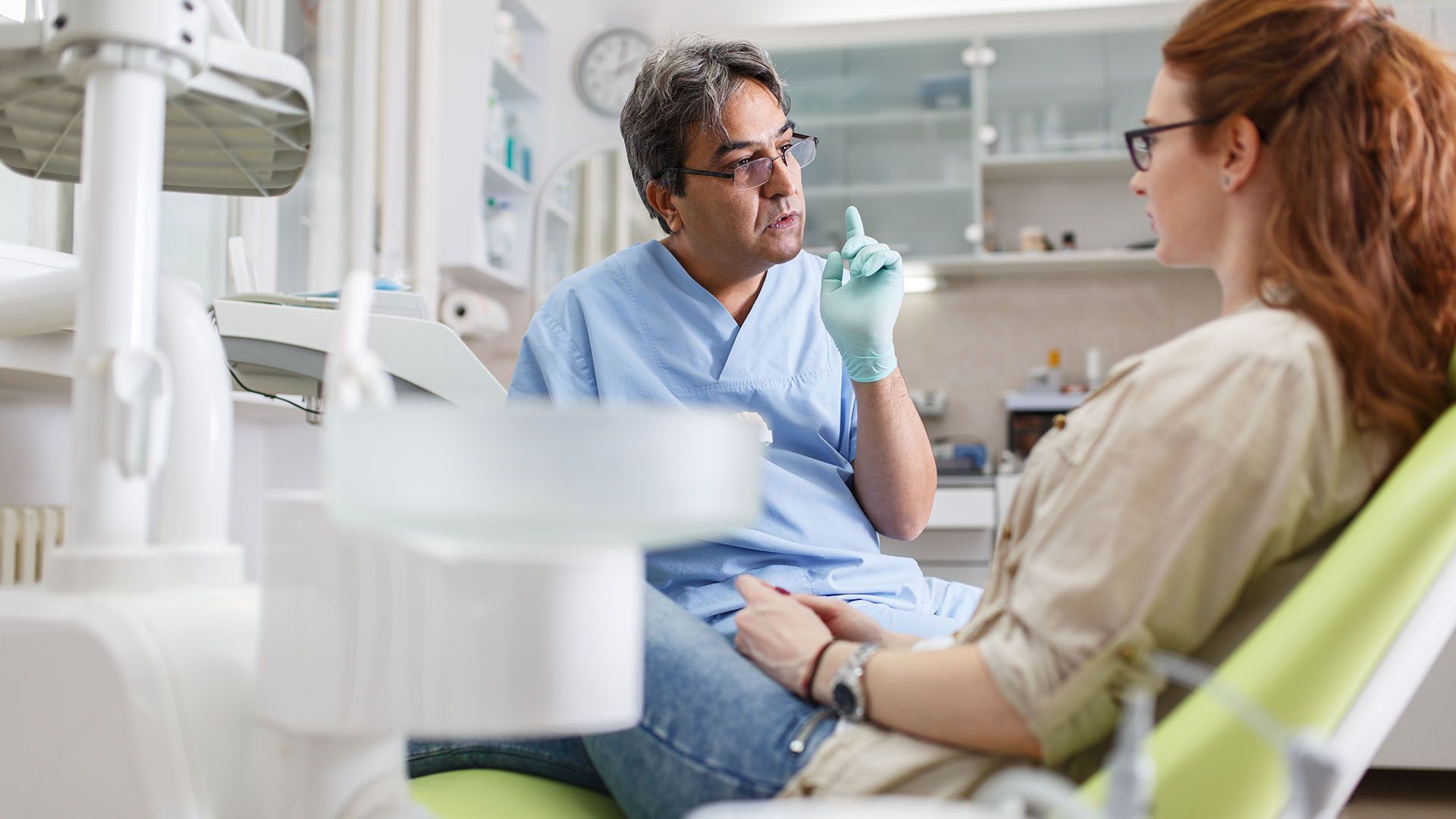 Dentist in blue scrubs points upwards, talking to a female patient in a dental chair.