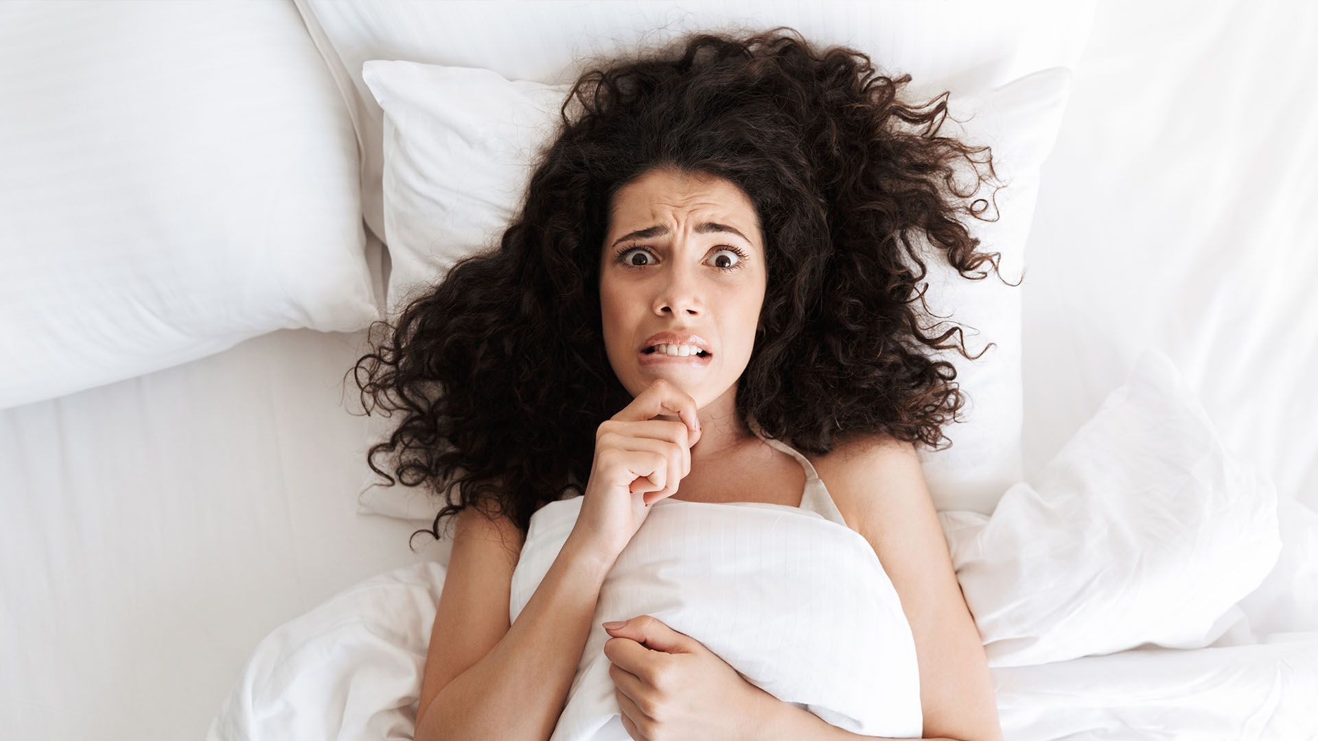Woman with curly dark hair in a bed, looking scared while clutching a pillow.