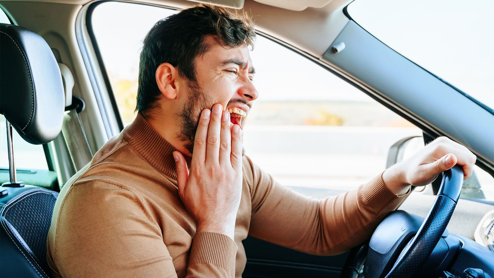 Man driving a car, grimacing and holding his jaw, likely experiencing tooth pain. Interior car setting.