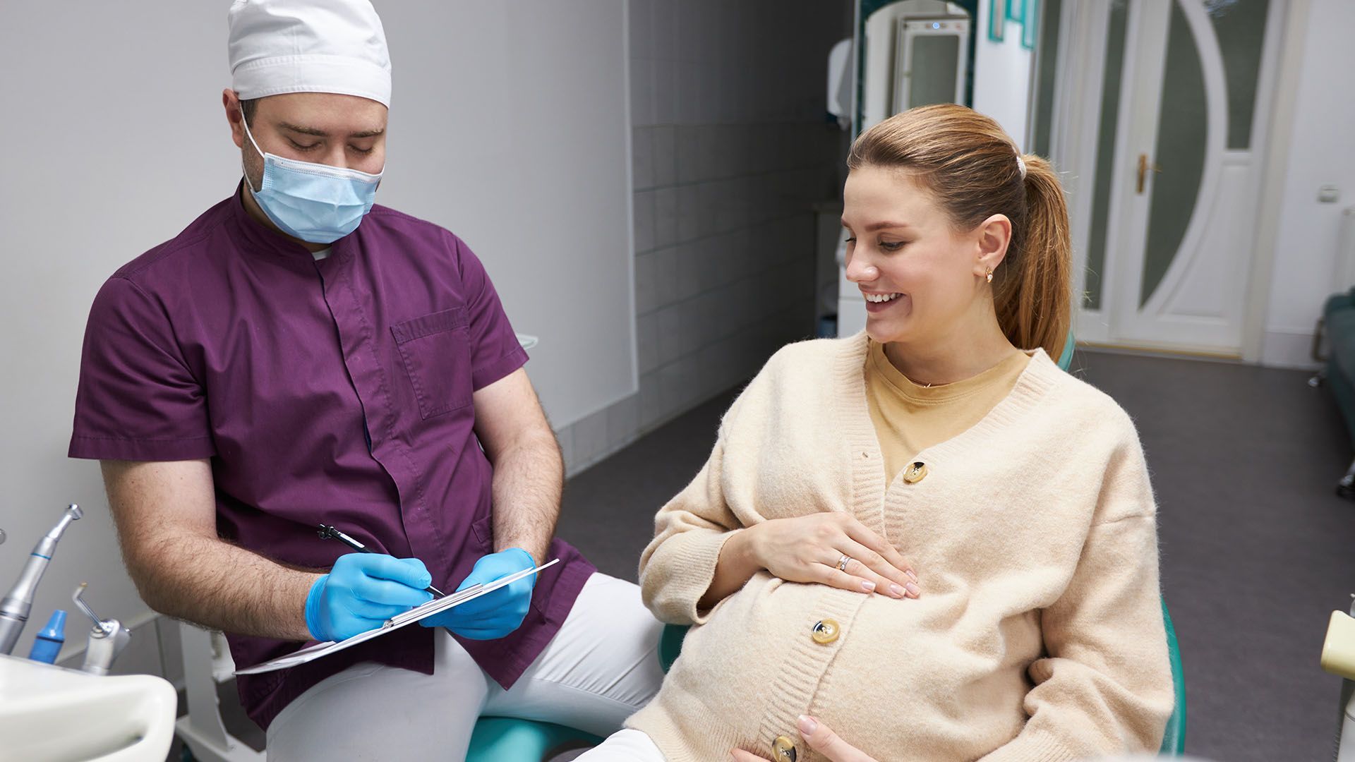 A pregnant woman smiles while sitting in a dental chair, talking to a dentist in a purple shirt and mask, who is taking notes.