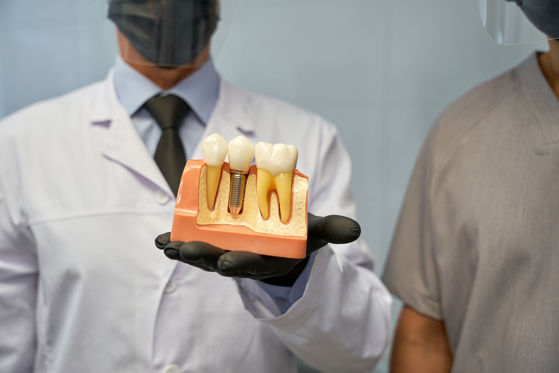 Person in lab coat holding a dental implant model next to a standing patient in a clinic