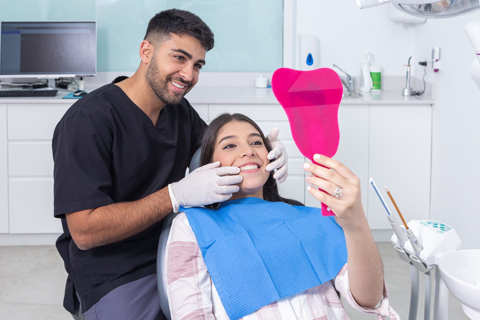 Dentist showing a patient her smile in a mirror. Bright dental office; dentist wears gloves and scrubs.