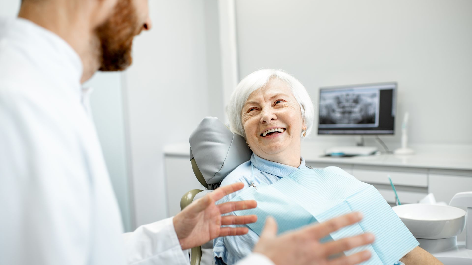 Dentist speaking to elderly woman in dental chair, both smiling. X-ray image visible in background.