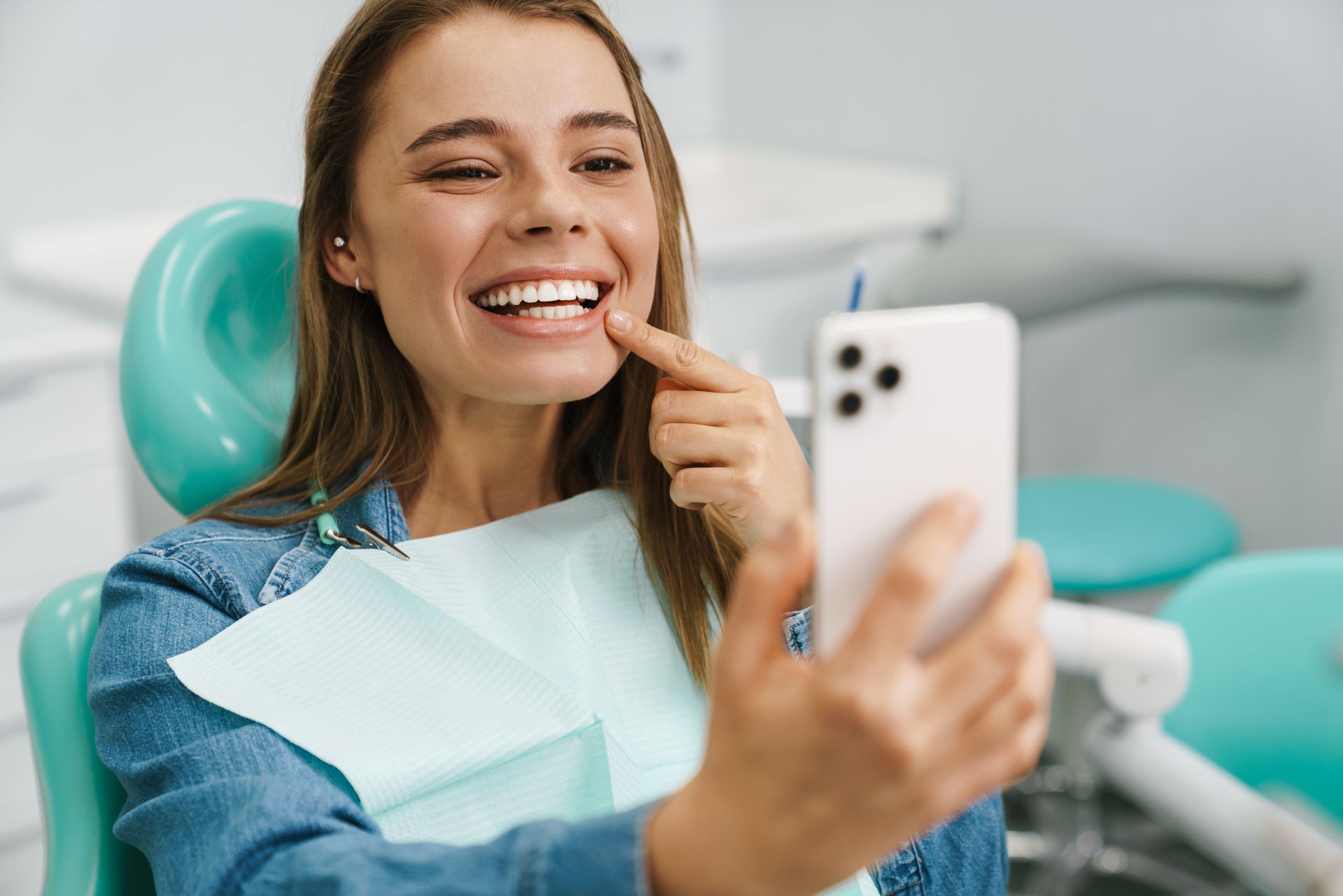 Woman smiling in a dentist chair, taking a selfie while wearing a bib and holding a toothbrush.