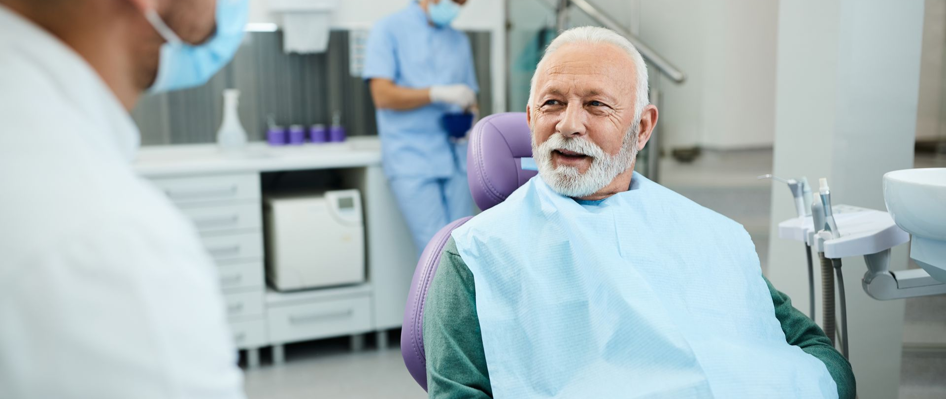 An older man in a dentist's chair, a bib on, talking to a dentist; another person in background.