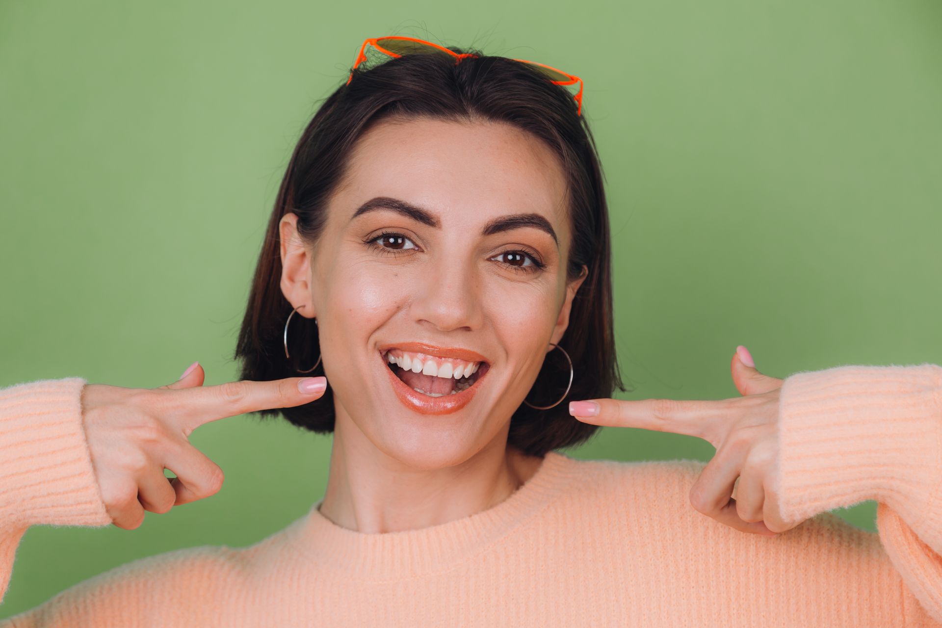 Smiling woman in a peach sweater pointing at her teeth against a green background