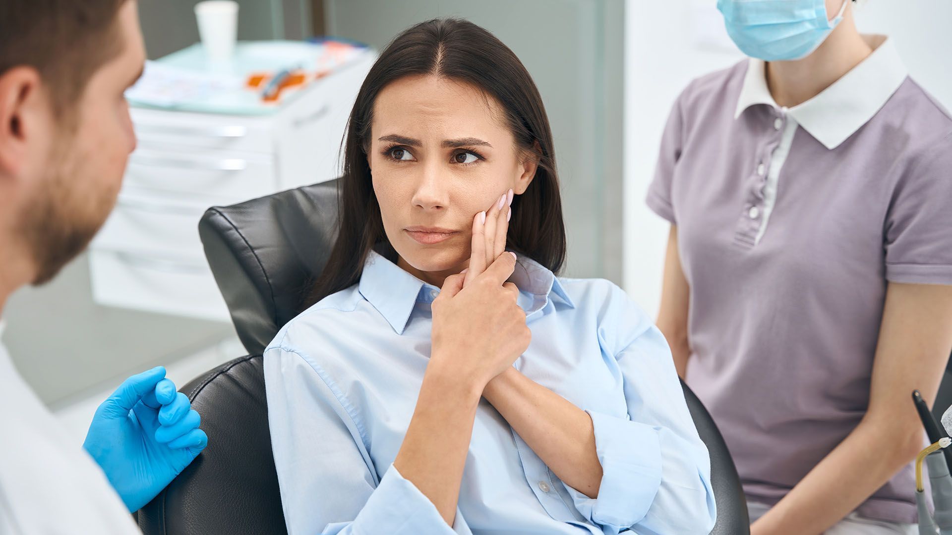 Woman in dental chair, holding cheek, appears distressed as dentist consults with assistant.