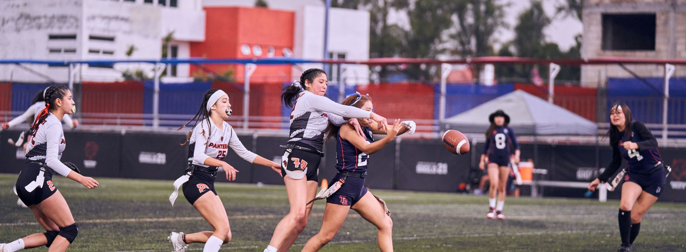 Flag football game at night, a player in red jersey runs with the ball as others pursue on a green field.