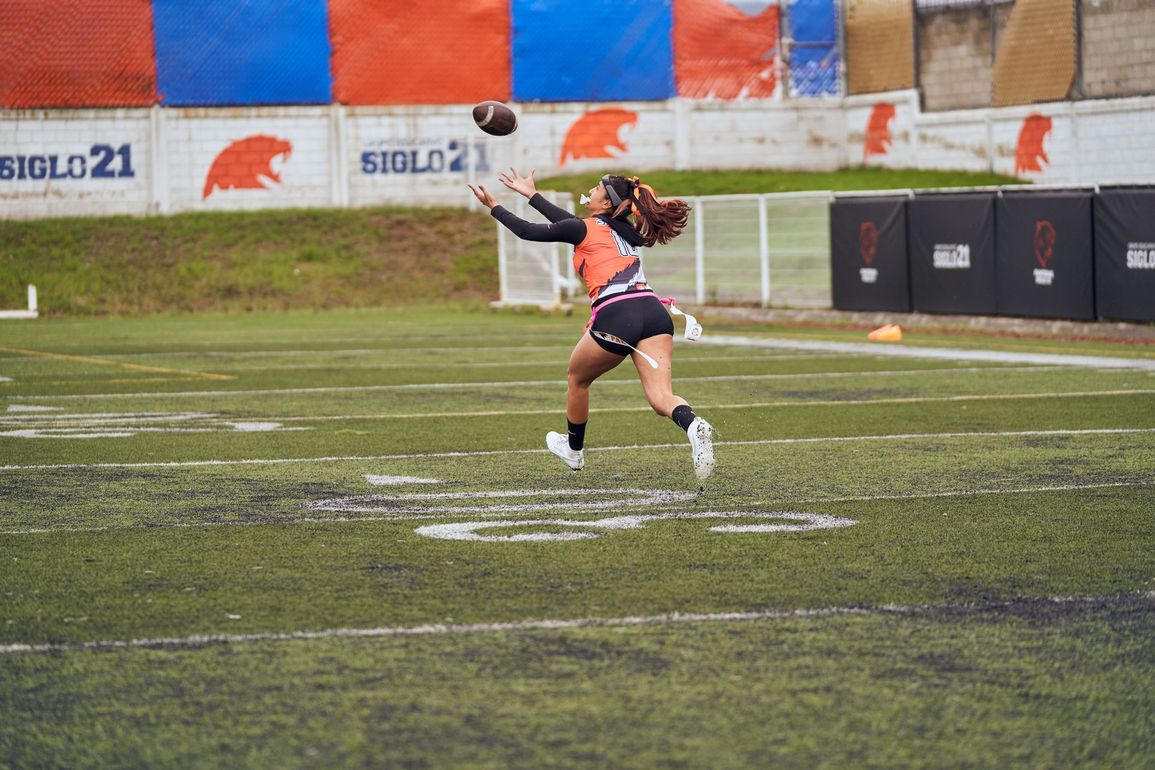 Football players in action on a grassy field; one runs with the ball, pursued by two opponents.