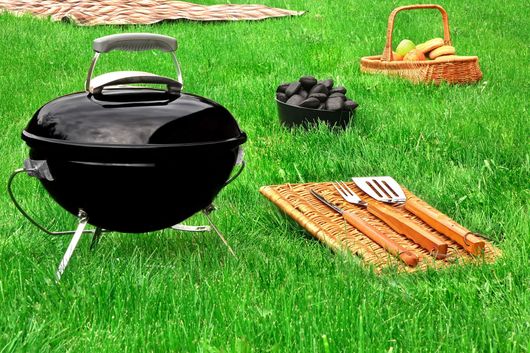Griglia a carbone nera con utensili, carbone e cestino da picnic su un prato verde all'aperto