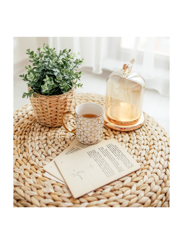 A cozy vignette: a plant, mug of tea, open book, and lit cloche on a woven table.