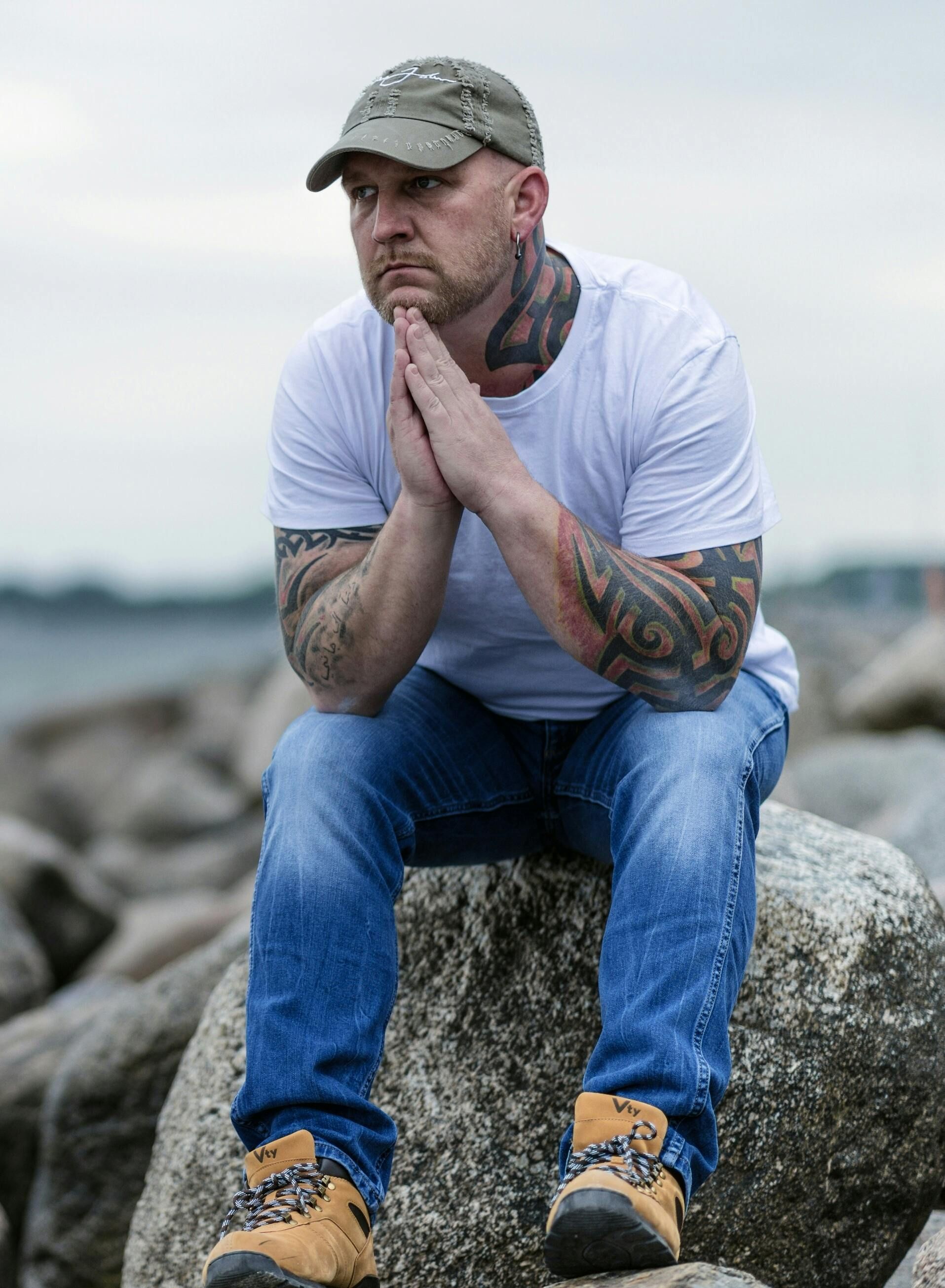 Man with tattoos sits on a rock, hands clasped, looking thoughtful, wearing a cap, jeans, and boots.– stress management