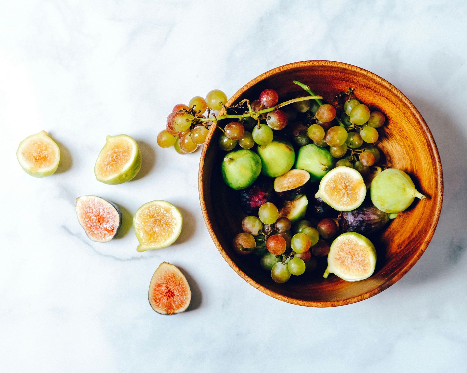 Wooden bowl of grapes and figs on a marble surface, with figs scattered around.– healthy recipes