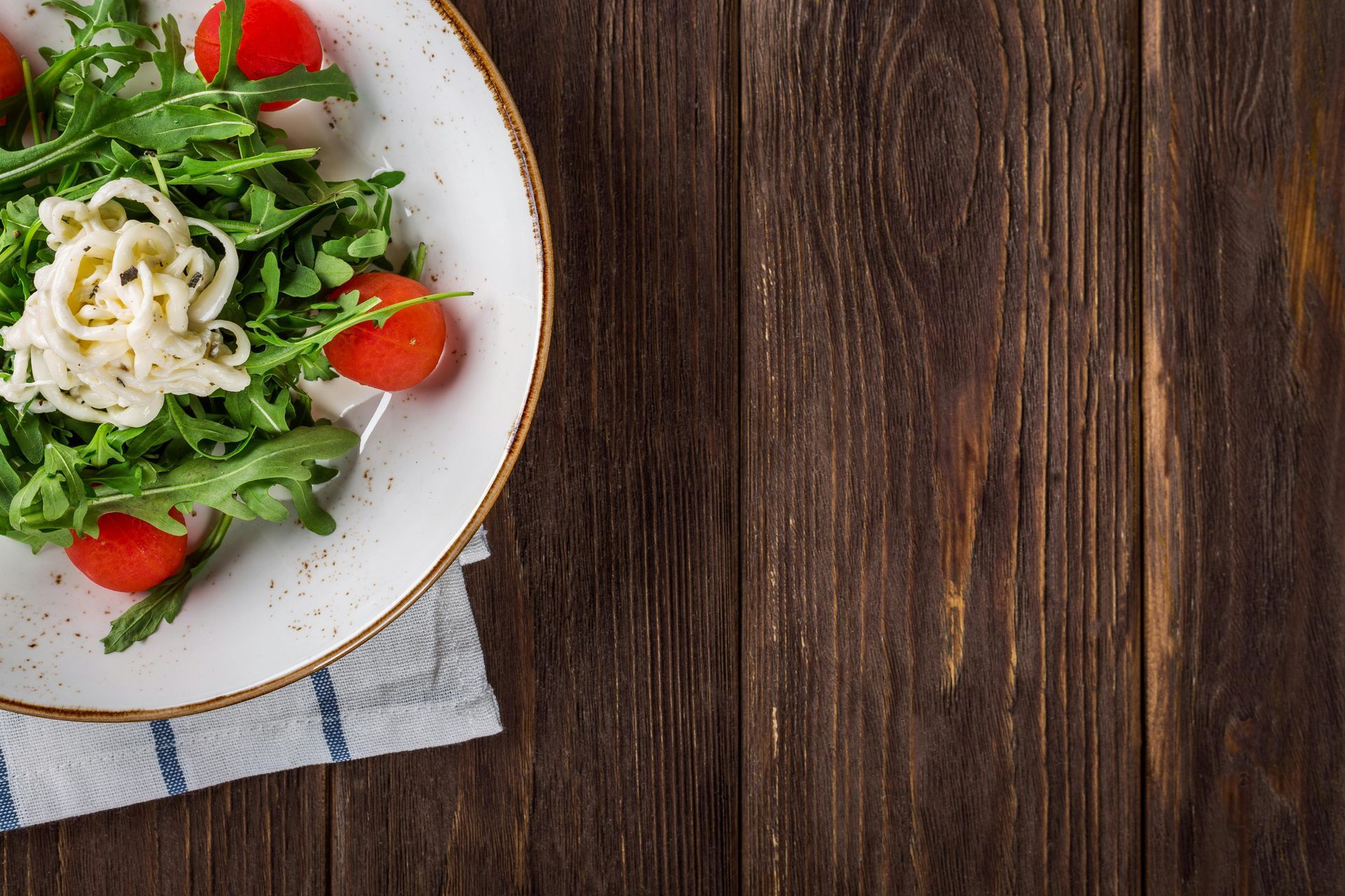 Salad on a plate with arugula, cherry tomatoes, and cheese, next to a wood table.