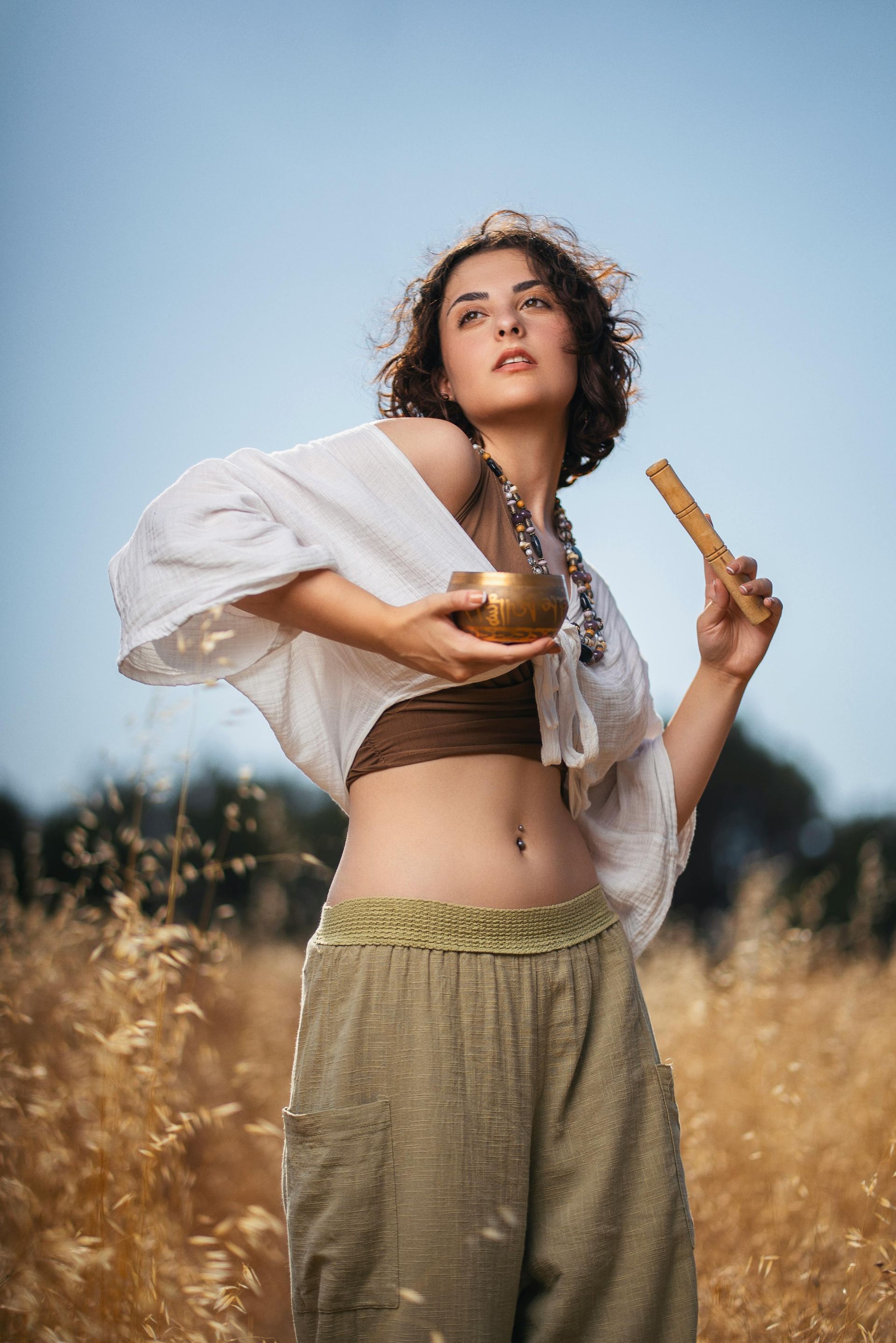 Woman in field holds a singing bowl and mallet, looking upward. Wheat field, light clothing.– self-care strategies