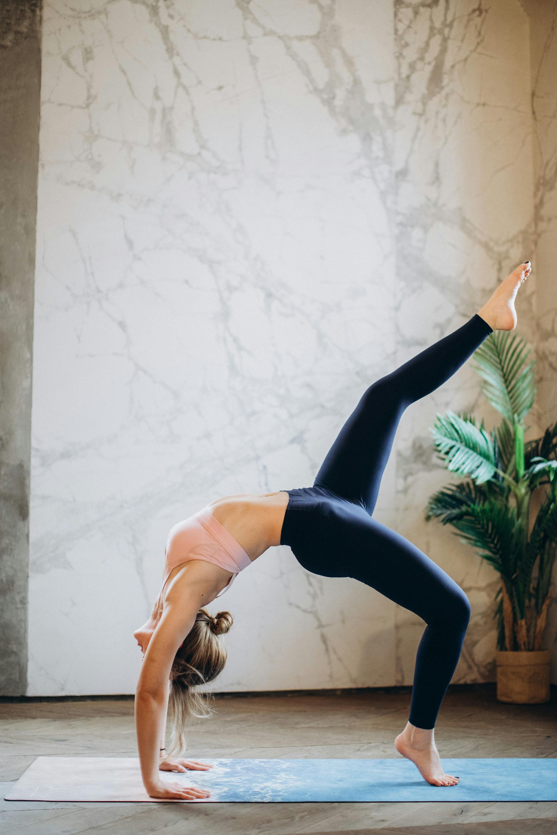 Woman in yoga wheel pose, lifting one leg, in a light-filled room with marble wall, plant, and yoga mat.