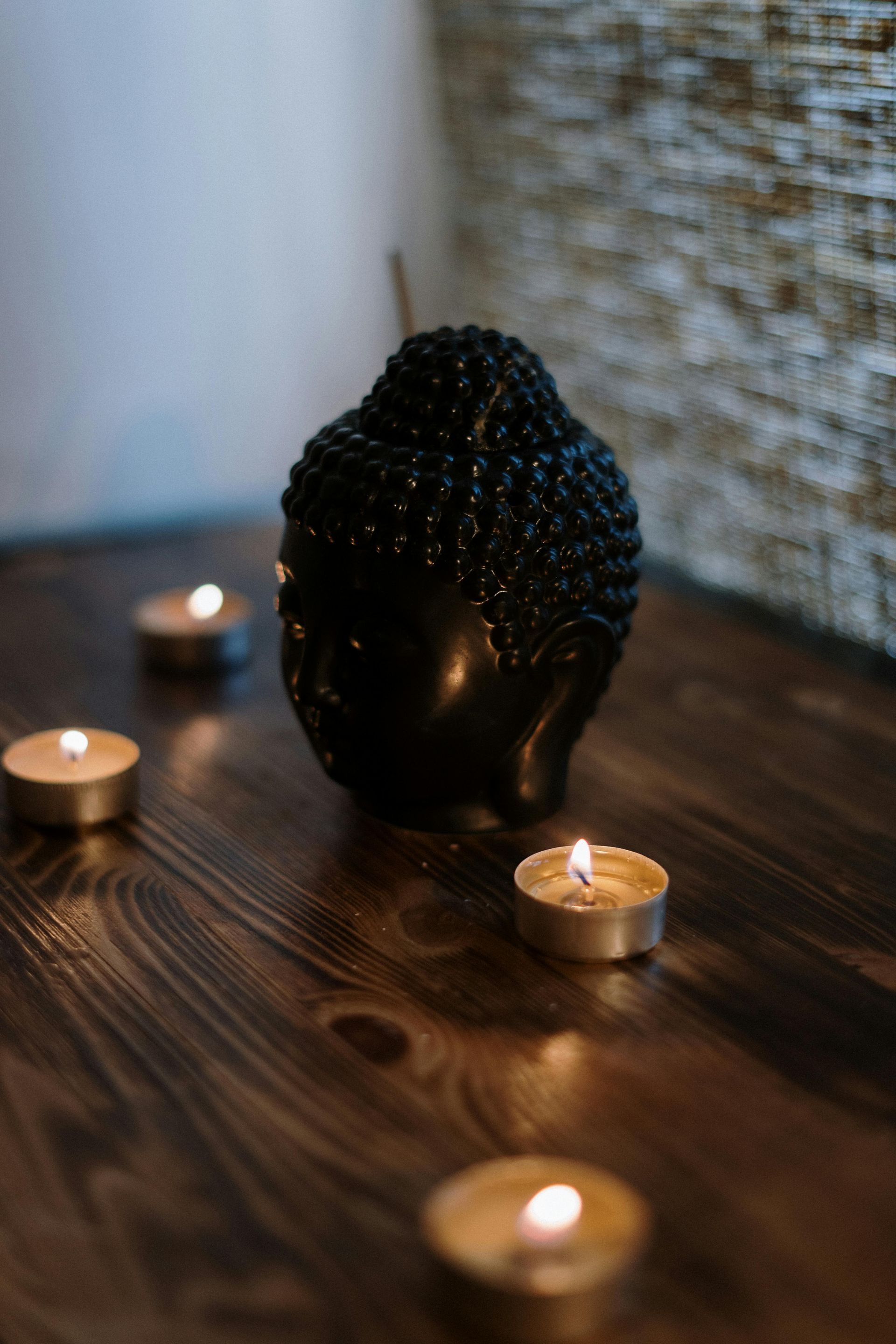 Black Buddha head statue with flickering candles on a wooden surface.– holistic health