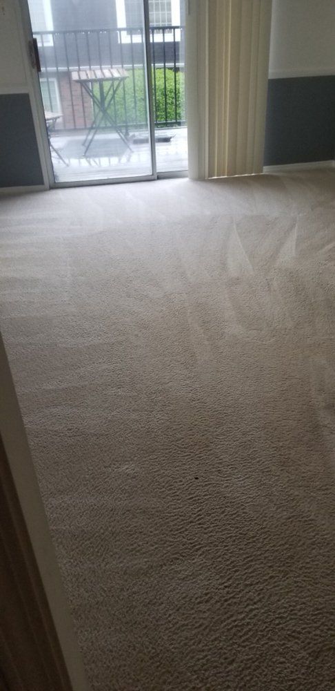 Empty beige carpeted room with sliding glass doors and curtains, leading to a balcony.