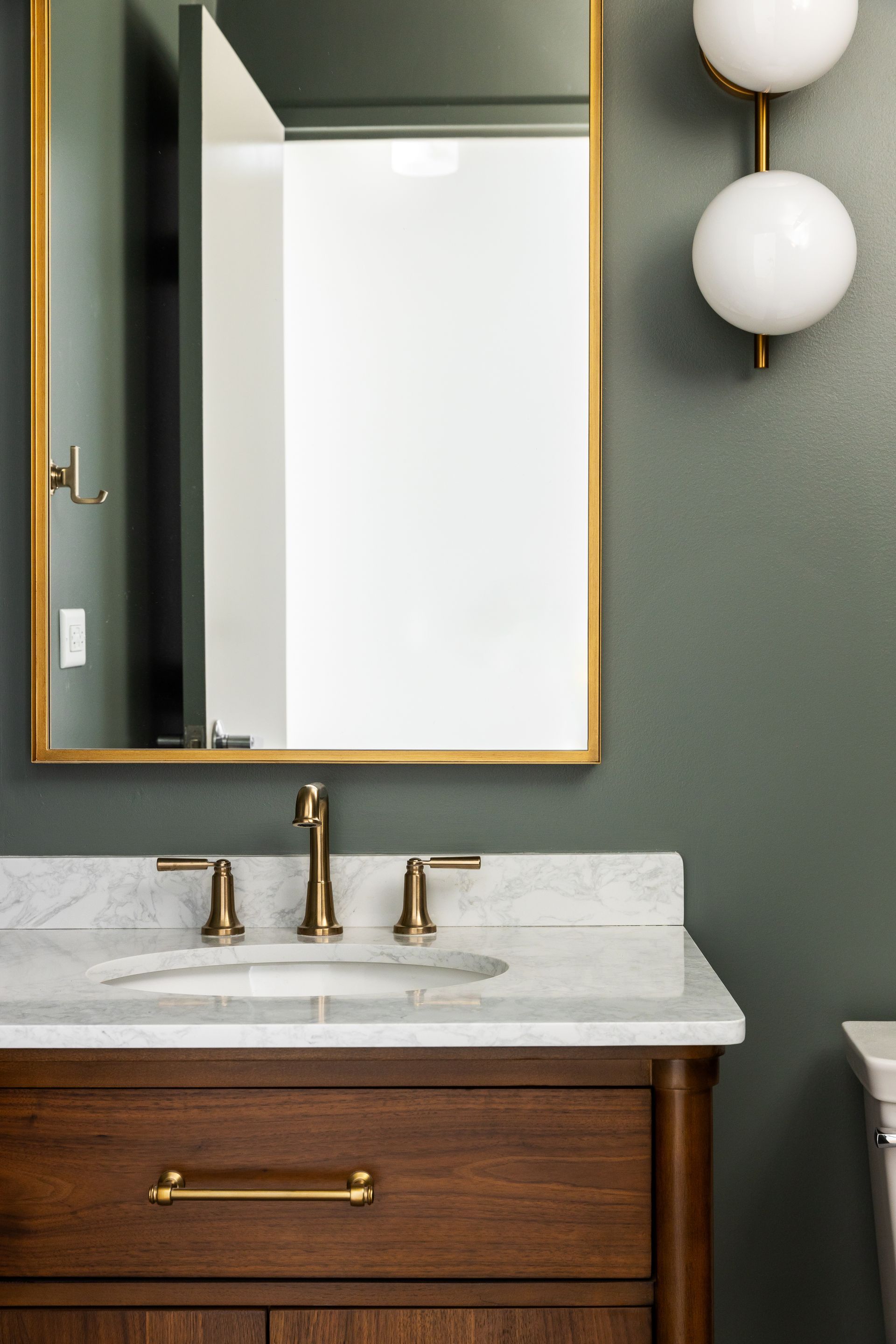 Bathroom vanity with gold-framed mirror, marble countertop, wooden cabinet, gold fixtures, and globe sconce on green wall.