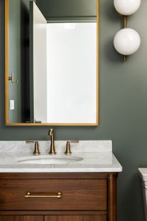 Bathroom vanity with a gold-framed mirror, marble countertop, wooden cabinet, and brass fixtures. Dark green wall.