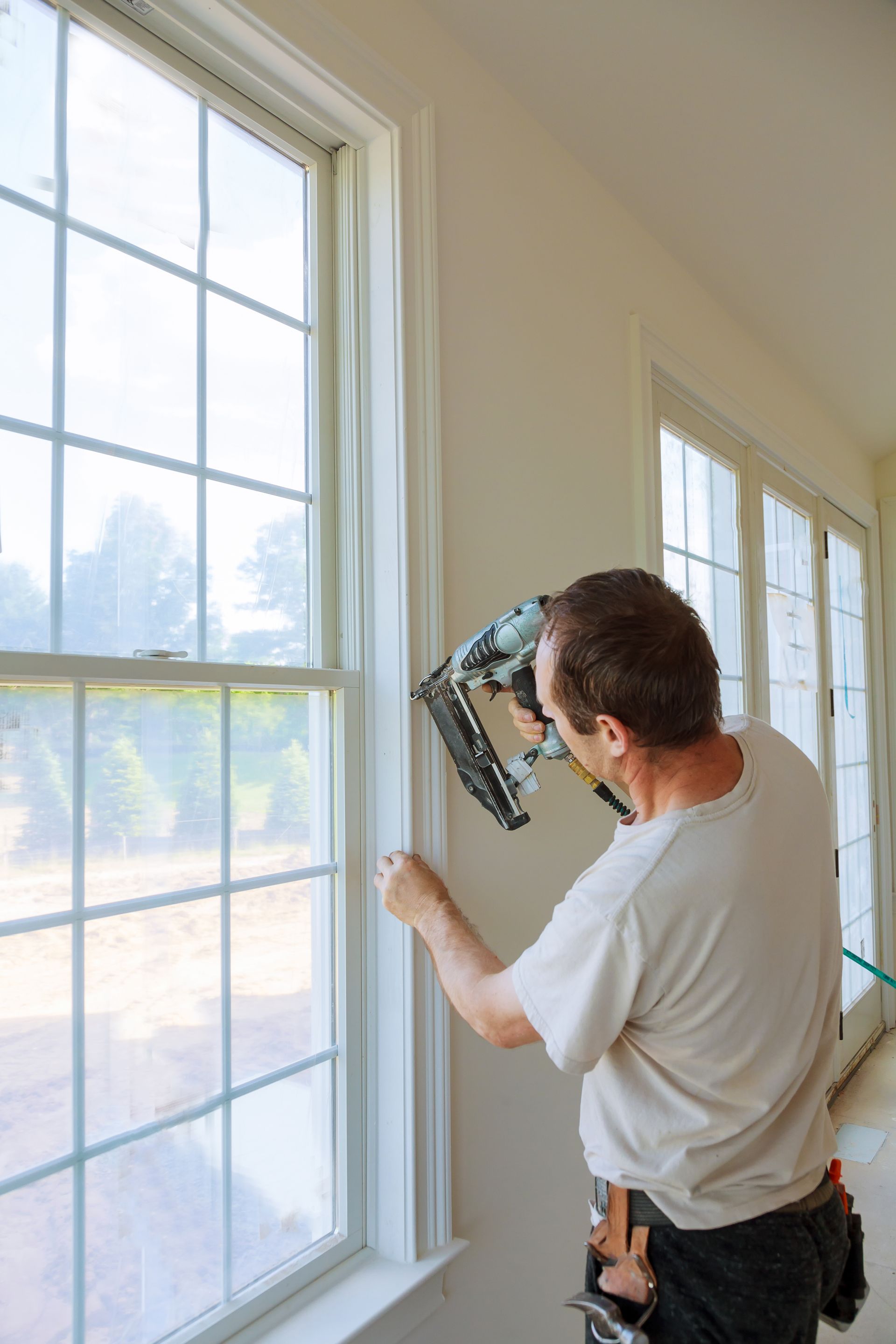 Man using a nail gun to install window trim in a room with white walls and windows.