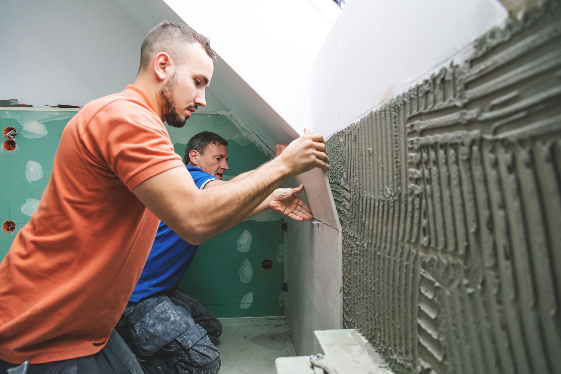 Two workers installing tiles on a wall in a room; one applies a tile.