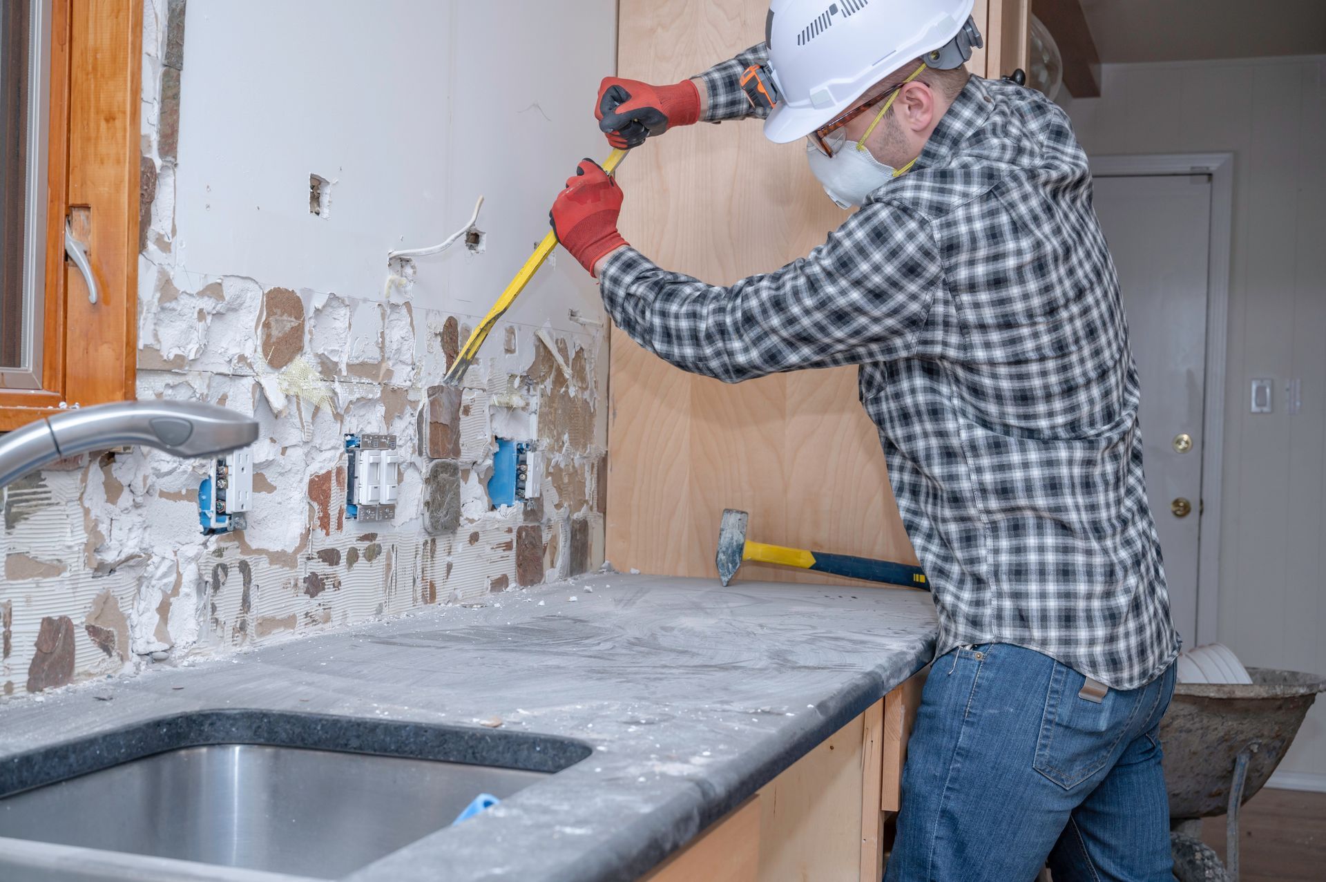 Construction worker in hard hat, mask, and gloves measuring wall near kitchen sink.