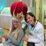 Two smiling women in a clinic wearing a giant red-and-gold crown and a lab coat