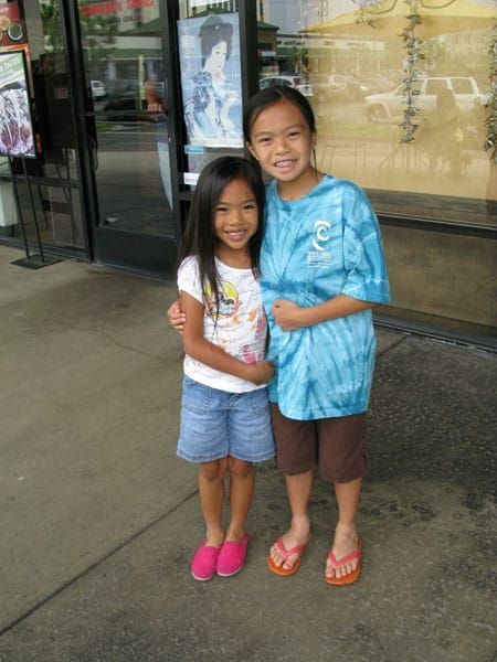 Two smiling children standing outside a store, holding each other and wearing bright summer clothes.