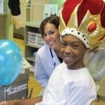 Woman smiles beside child wearing a red-and-gold crown beside a blue balloon in a classroom