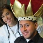 Two smiling people wearing festive crowns pose indoors against a red curtain.