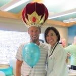 Two smiling people in a room, one wearing a crown-shaped balloon hat and holding a blue balloon.