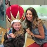 Girl in a large red crown smiling with a woman beside her indoors.