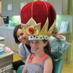 Child wearing a large red and gold crown while smiling, with another person standing behind in a room