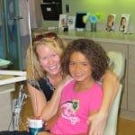 Two smiling children sit together indoors, one in a pink shirt beside another in black.