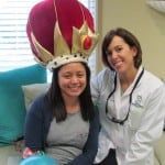 Smiling woman wearing a large crown beside a woman in a white coat, seated indoors.