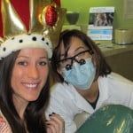 Two friends in festive hats posing together, one wearing a mask, holding a green balloon indoors