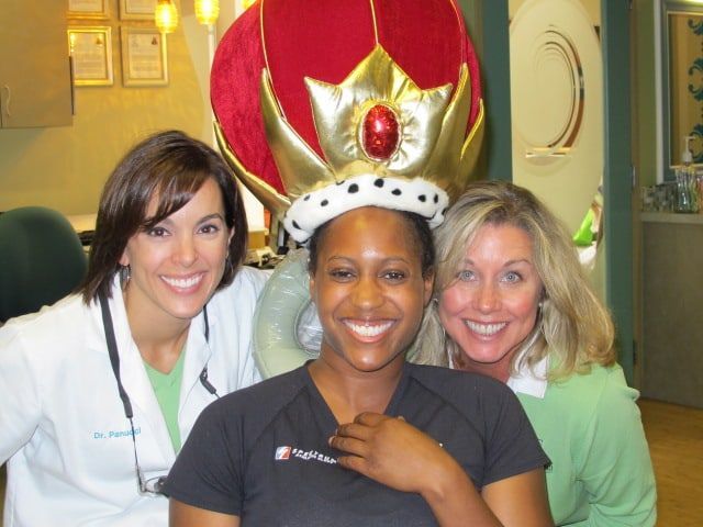 Three smiling people pose indoors, with one wearing a red-and-gold crown.