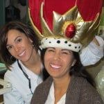 Two smiling people posing, one wearing a large red-and-gold crown and white fur-trimmed costume.
