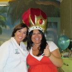 Two women smiling, one wearing a red crown, posing at an indoor celebration.