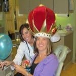 Smiling woman in a large red and gold crown, seated in a decorated room with another person nearby