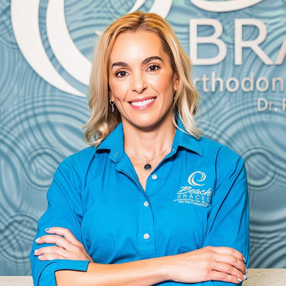 Smiling woman in a blue shirt with crossed arms, standing before an orthodontics office backdrop
