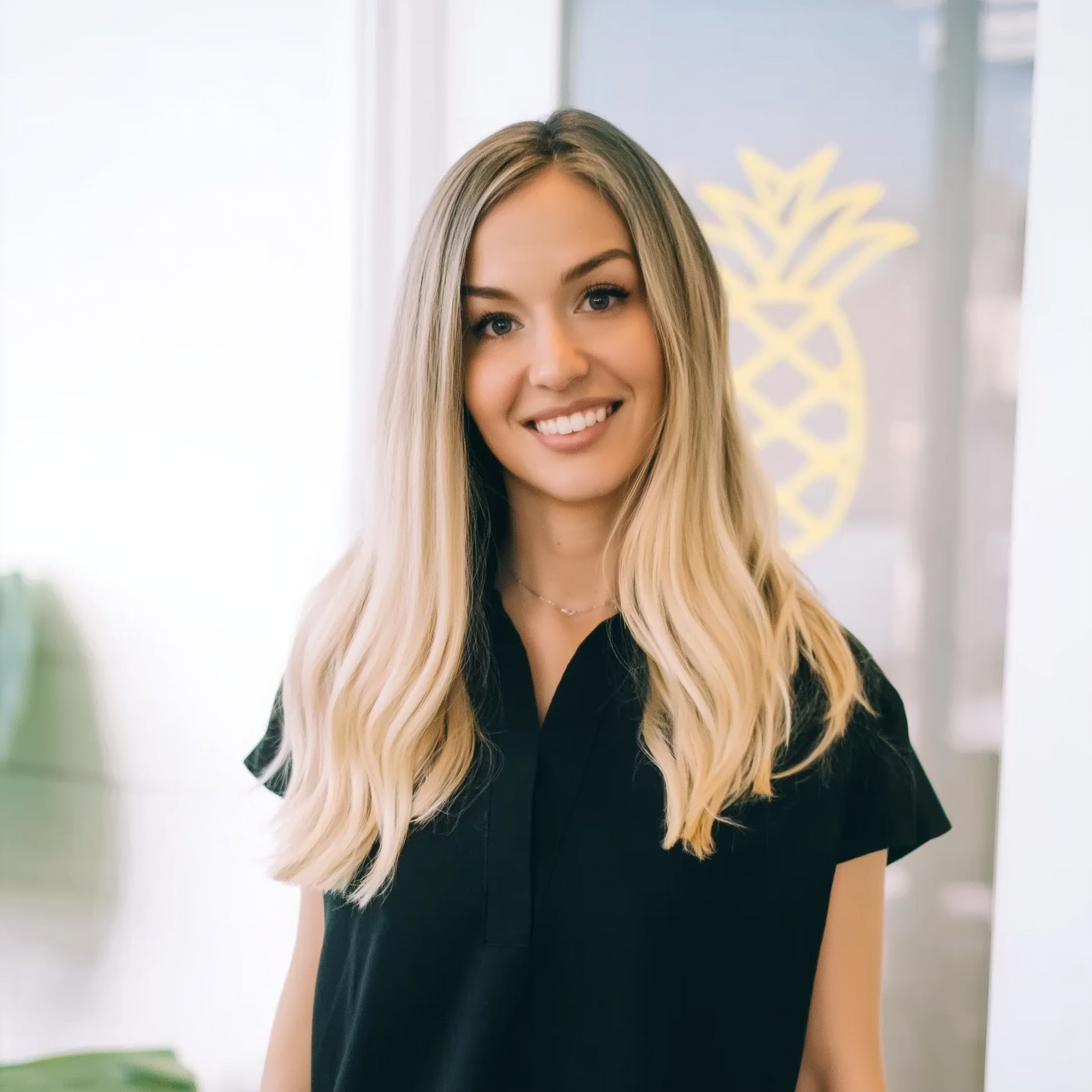 Smiling woman in a black shirt standing indoors with a bright pineapple logo in the background
