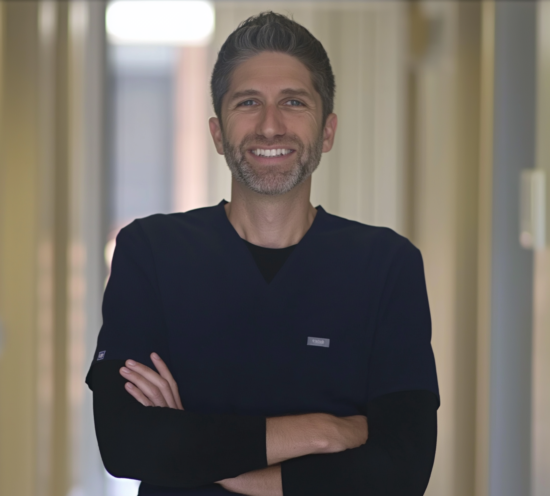 Smiling person in a dark shirt standing in a hallway with arms crossed