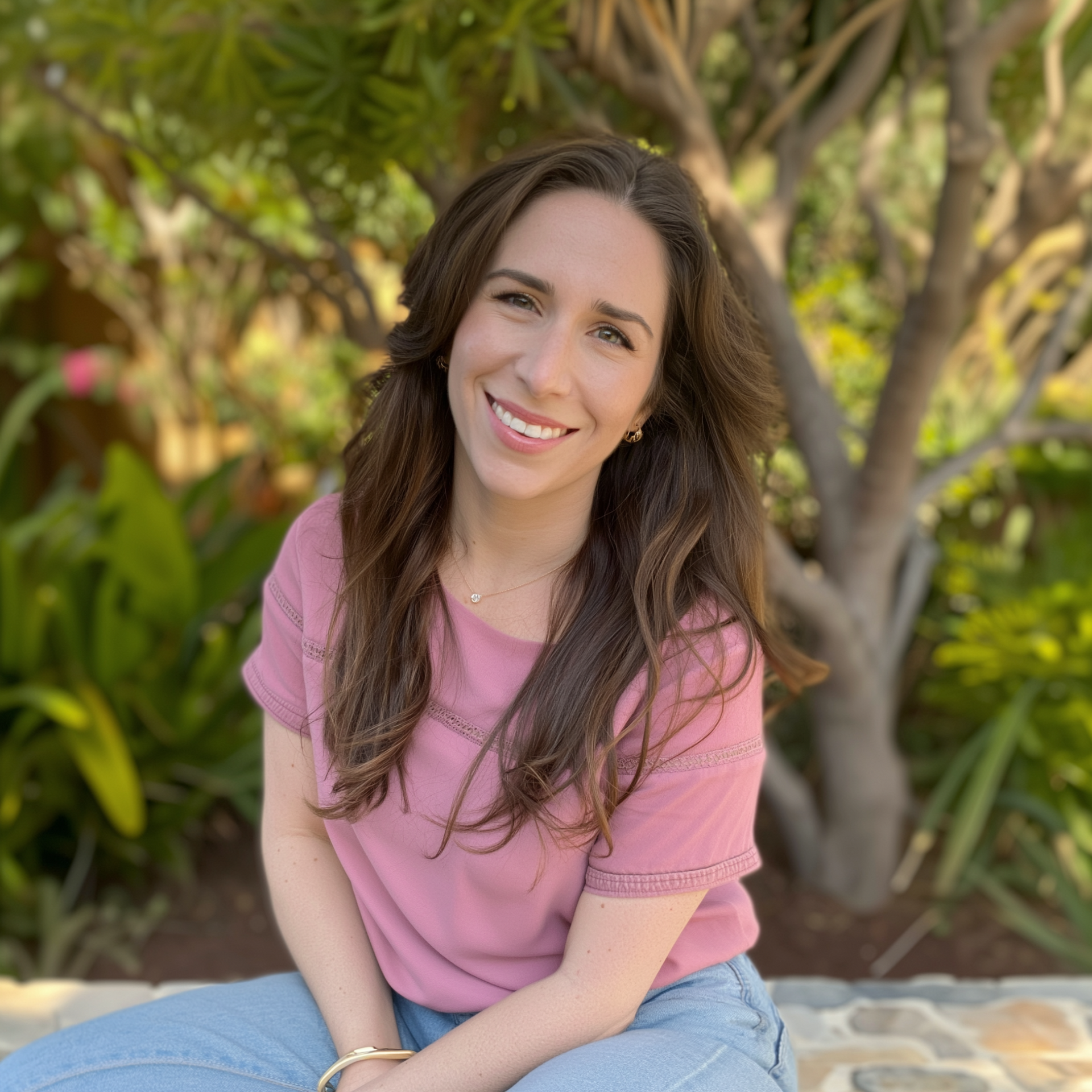 Smiling woman in a pink shirt sitting outdoors in a leafy garden